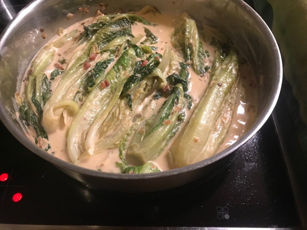 Foto vom fertigen Gericht. In der Pfanne die gedünsteten Salatköpfe in einer Senf-Sahne-Soße mit knusprigen Speckwürfeln. Die Pfanne steht noch auf dem Herd.