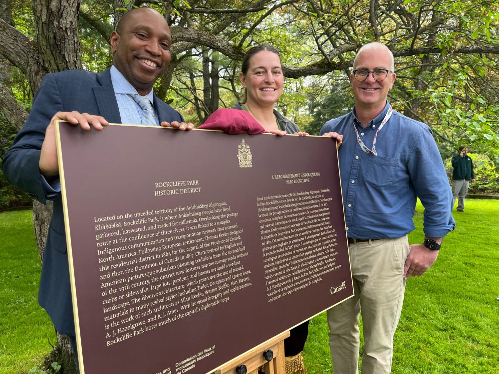 Rawlson, Stéphanie and Jeff pose around an historical designation plaque outside on a park lawn with trees in the background.