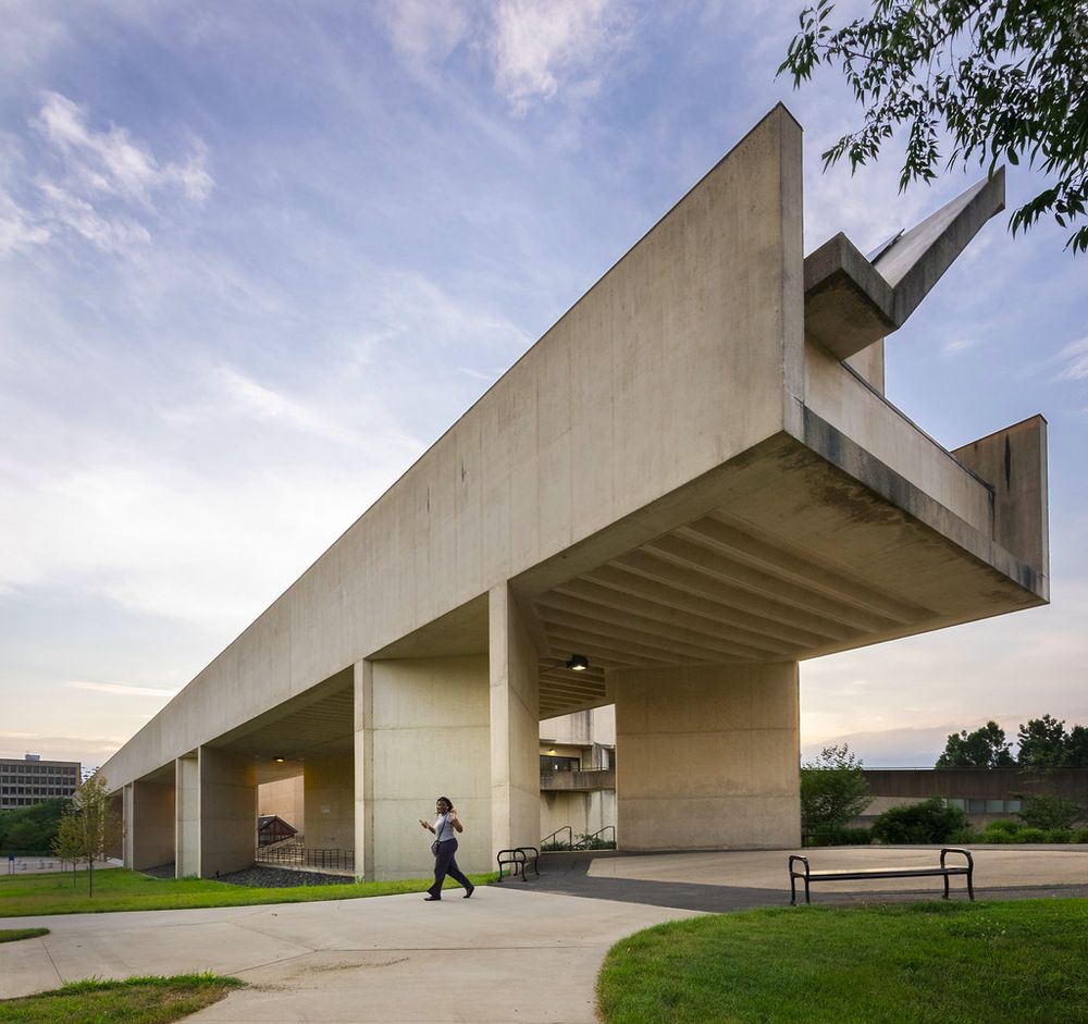 Photograph by Darren Bradley of the UMass Amherst Fine Arts Center, which he describes as "the most impressive and interesting" example of modernist architecture on the campus. The photo, which is the featured image for Bradley's post, "UMASS Amherst: The Reluctant Modernists," shows one end of the building, a large concrete structure with bold angles that juts into space like an unfinished bridge. A student is passing under the suspended portion of the structure on a broad sidewalk that forks in multiple directions, with green lawn on all sides. Another modernist structure is visible in the background and the whole scene is framed against a blue sky with wispy clouds.