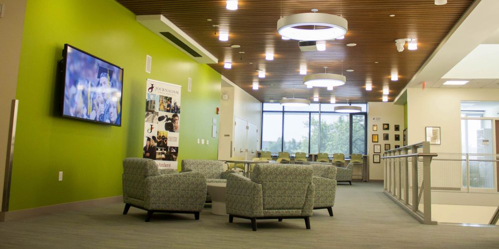 Interior view of the Journalism Department's common space inside the Integrative Learning Center at UMass Amherst. Cozy pods of chairs are flanked by digital signage and an interior staircase. The view of campus is visible through the windows in the background.