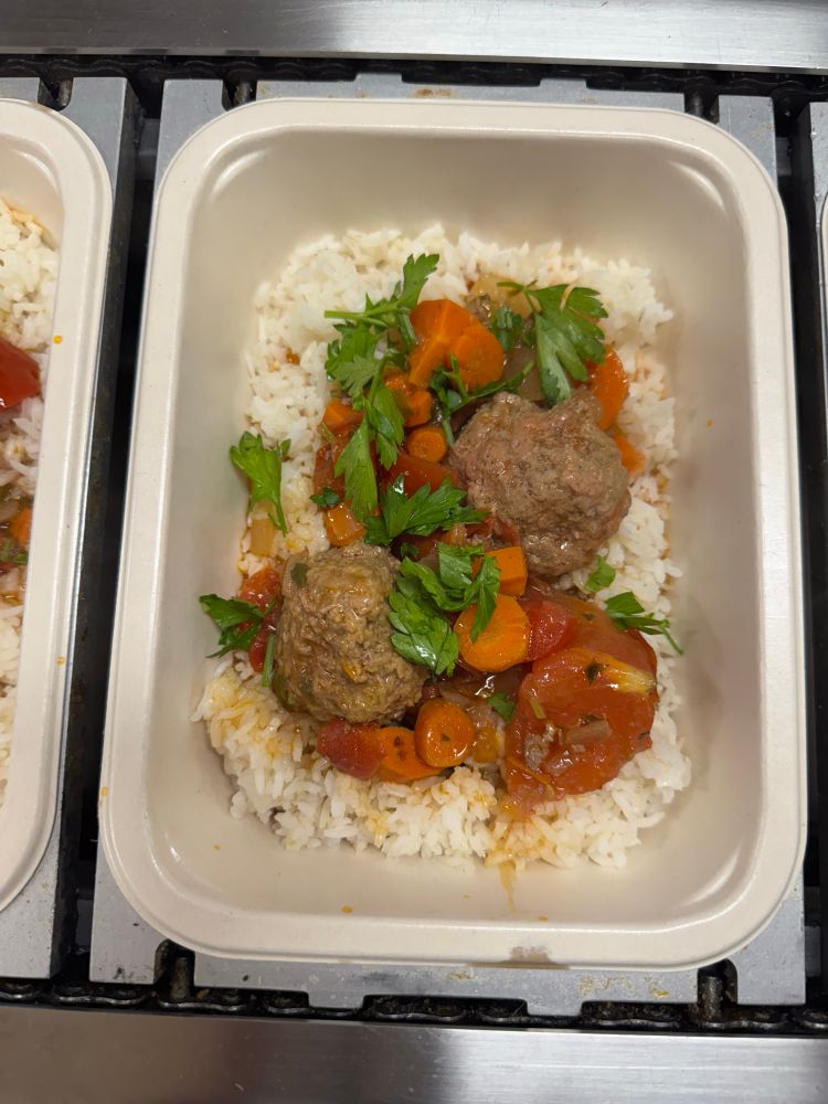 Photo of a compostable tray filled with one meal of white rice, big juicy meatballs, and a sauce with carrots and tomatoes. Cilantro over the top adds a pop of vivid green