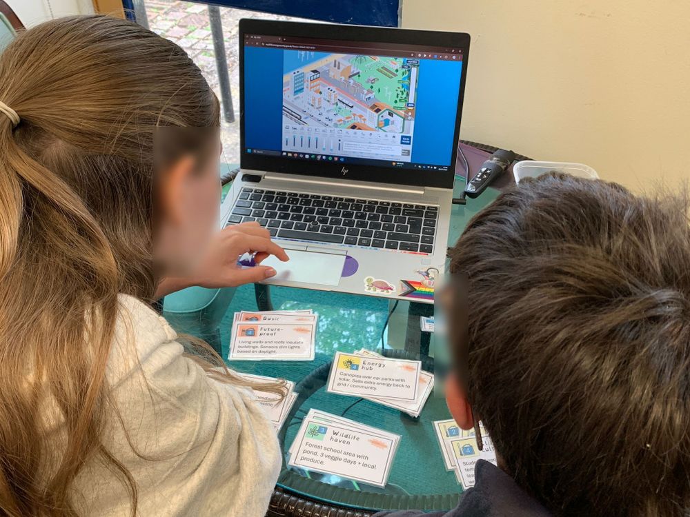 Two children sit at a table, looking at a laptop screen displaying a colorful game (BEIS My 2025 simulation) involving buildings and net zero actions. Several cards with text and images are spread out on the table, appearing to relate to the game.