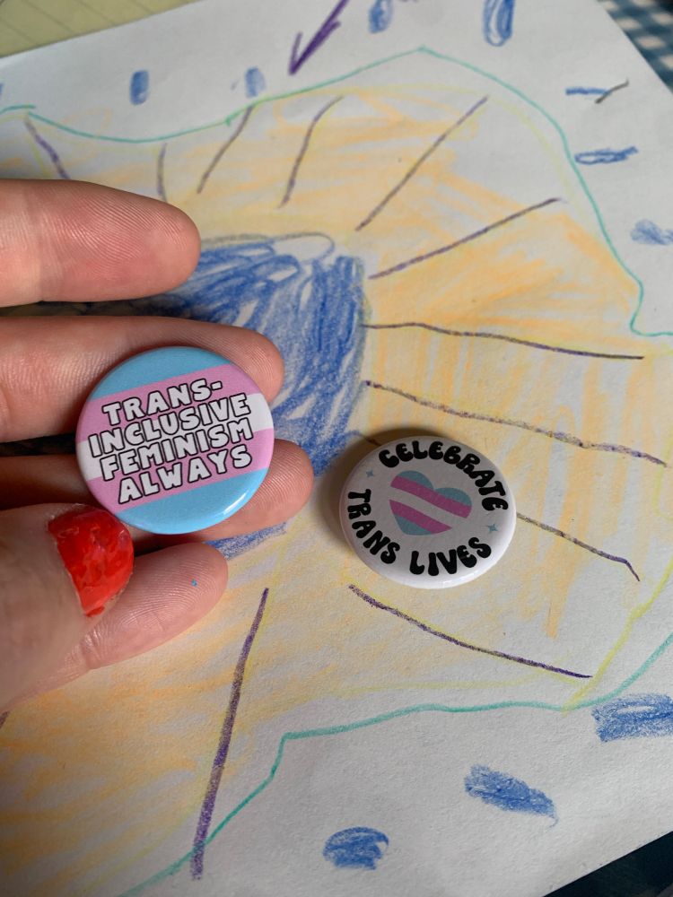 Two badges/pins that say “Trans Inclusive Feminism Always” and “Celebrate Trans Lives”