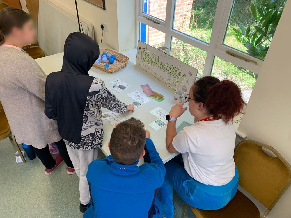 Four people gather around a table indoors, which is covered with educational materials including a 'Biodiversity' sign, informational sheets or cards, and a basket containing blue plastic eggs. Large windows in the background let in natural light.