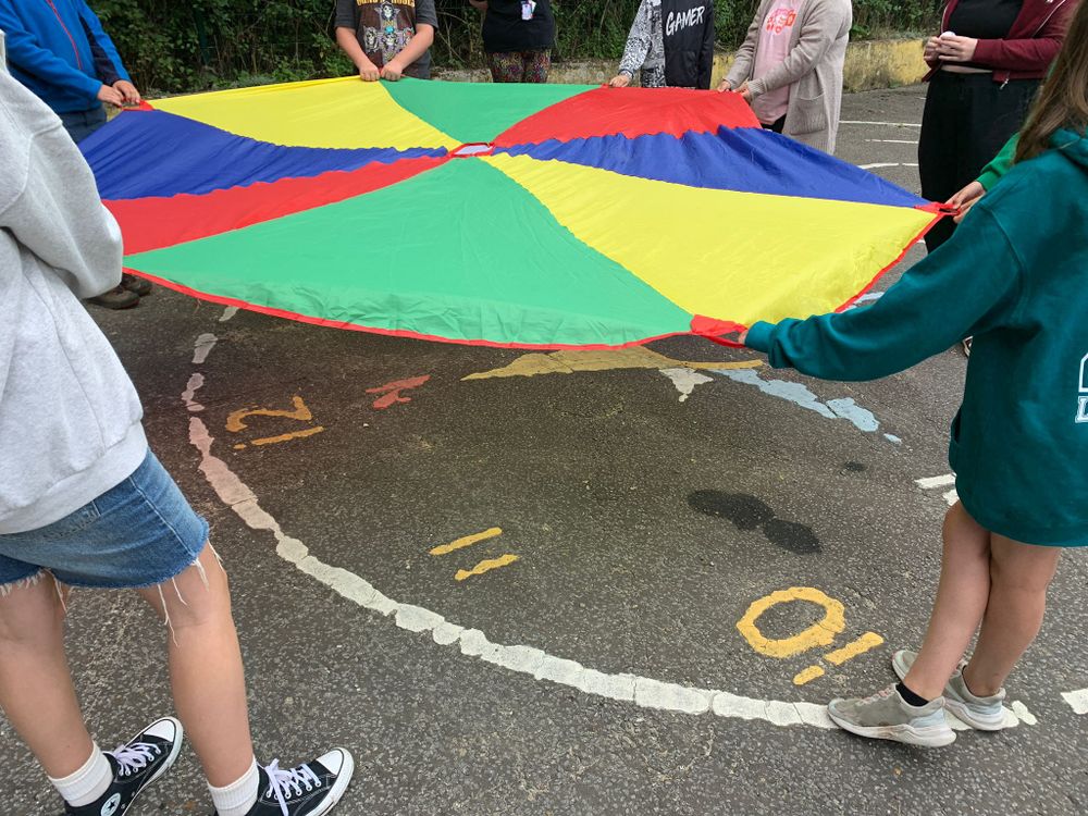 A group of children stand in a circle, each holding the edge of a colorful parachute divided into red, blue, green, and yellow sections. The ground beneath them features markings resembling a clock face, with numbers 10, 11, and 12 visible.