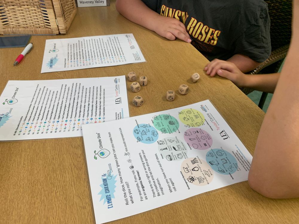 Two children sit at a table with educational materials, including two illustrated sheets titled 'Climate Education Game Pack,' wooden dice with symbols, and a red pen. The activity involves rolling dice and using cards to talk about green jobs.