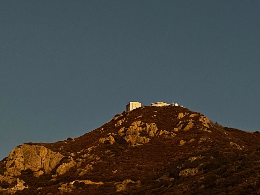 Grey-blue sky behind the summit of Mt Hopkins. The telescope and another building can be seen on top of the mountain. 