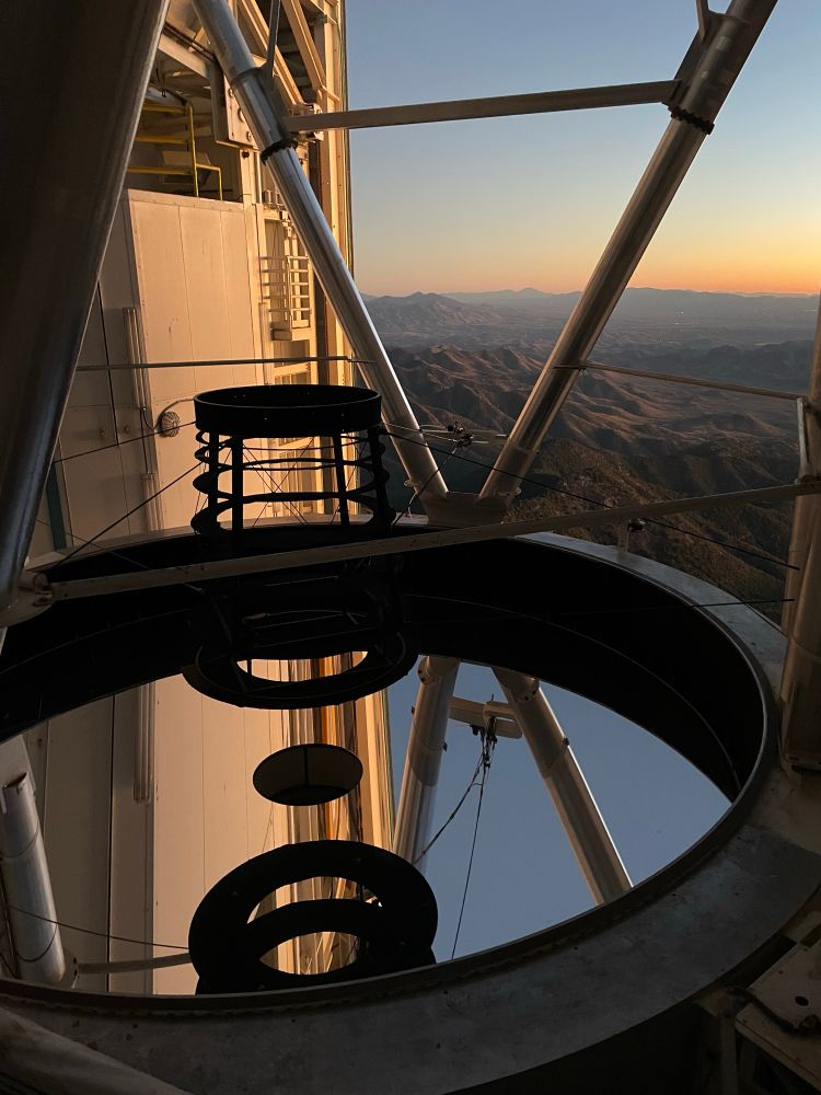 The MMT telescope as seen from the fourth floor of the chamber. Parts of the secondary support structure and the chamber wall can been seen reflected in the mirror. 
Mountains and a yellow/orange sky can be seen through the open chamber doors.