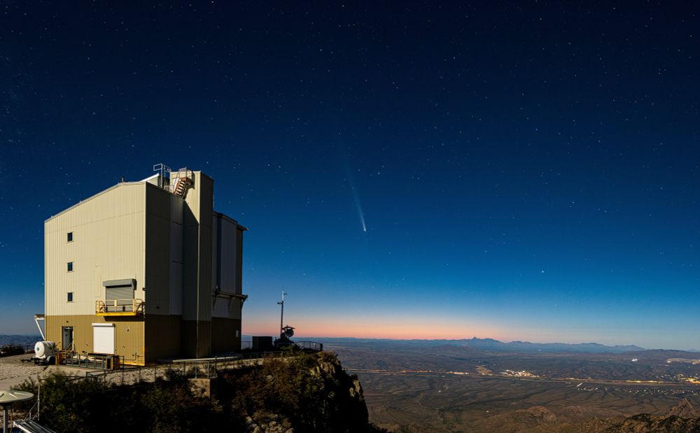 Comet C/2023 A3 (Tsuchinshan–ATLAS) is seen against a dark blue background with a slight orange sunset glow below. The MMT Telescope building can be seen in the foreground.