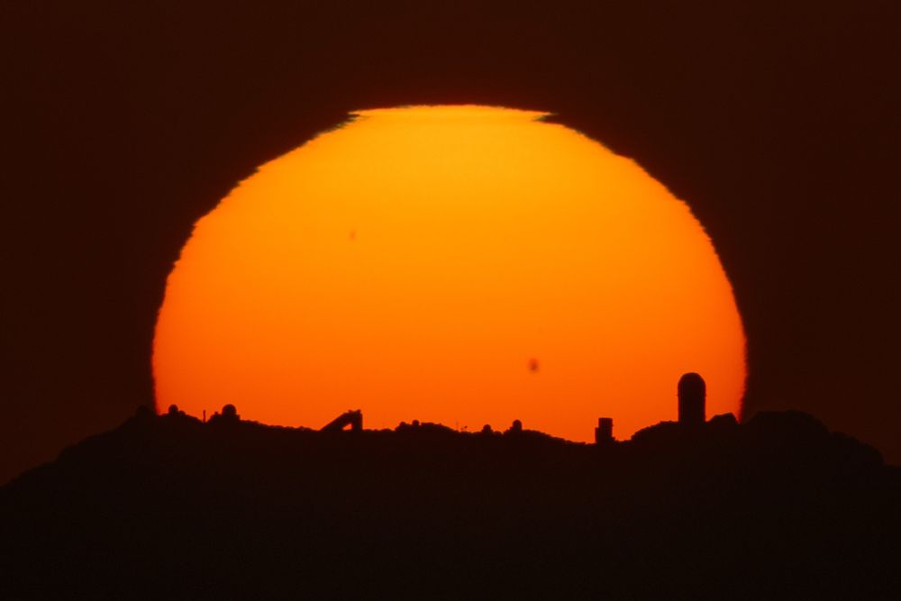 The sun setting behind Kitt Peak. A number of observatory buildings can be seen in the foreground, silhouetted by the sun. 