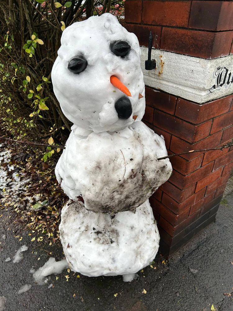 A snow person has two balls for body, a ball for head, a carrot nose, black objects for eyes and mouth. About 1m standing at the entrance to an alleyway