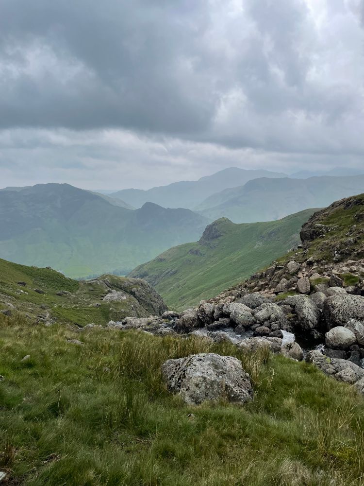 Picture shows grass in the foreground, a rocky stream going down, then green grassy ridges receding away into the background