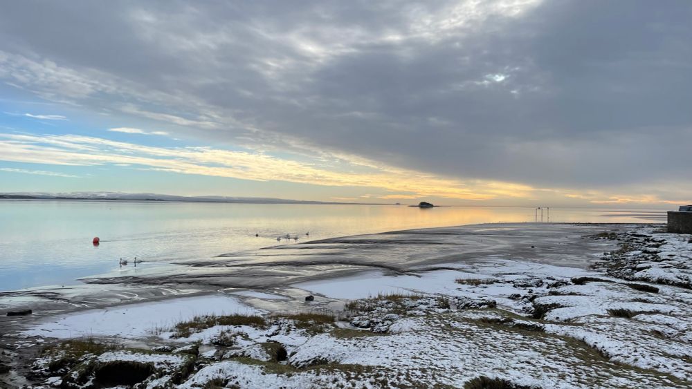 Colour photograph shows a landscape: in the middle of the image, at the horizon, from left to right, low grey and white smudgy looking hills, then a dark oval shape, an island and its reflection; in the sky above, from left to right, blue sky and white clouds, then darker clouds; in the foreground, from left, a blue and silvery water surface, then green and white snow covered grass land