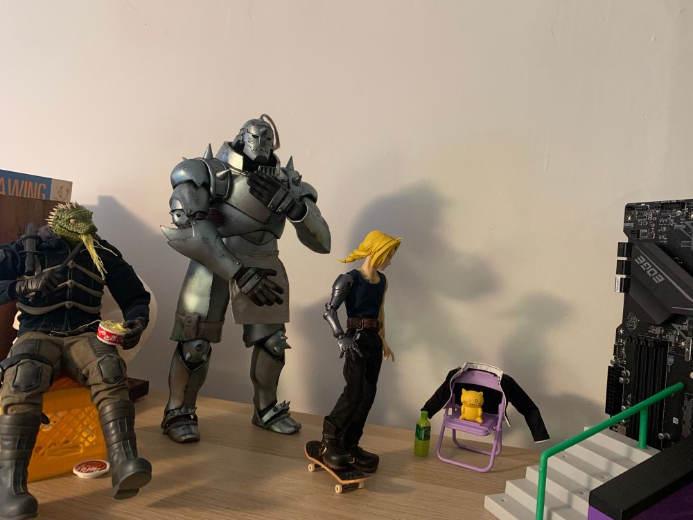 More posed figures. A scene in which Caiman, eating noodles with a knife, watches Ed Elric, who has a skateboard and is sizing up the stairs and railing in front of him, clearly preparing for a trick. Al stands behind his brother, looking worried. 