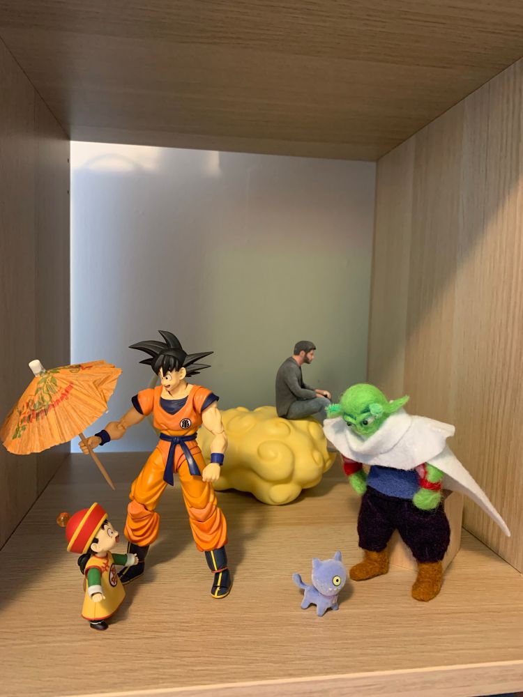 Posed figurines. On the left, Goku holds an orange umbrella over child Gohan. On the right, a handmade Piccolo looks at a miniature flocked Ugly Doll cat. In the background, Sad Keanu sits on Kinto’un.