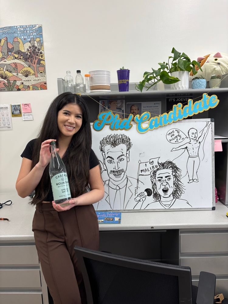 Tiffany, holding a bottle of fancy water, in front of a sign that says PhD candidate. 