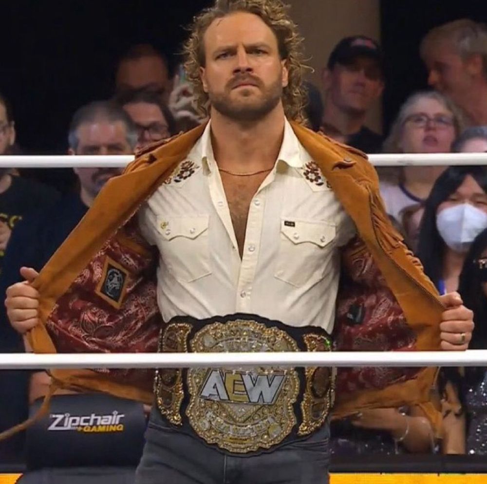 Hangman Adam page opening his jacket to show the AEW men;s title belt