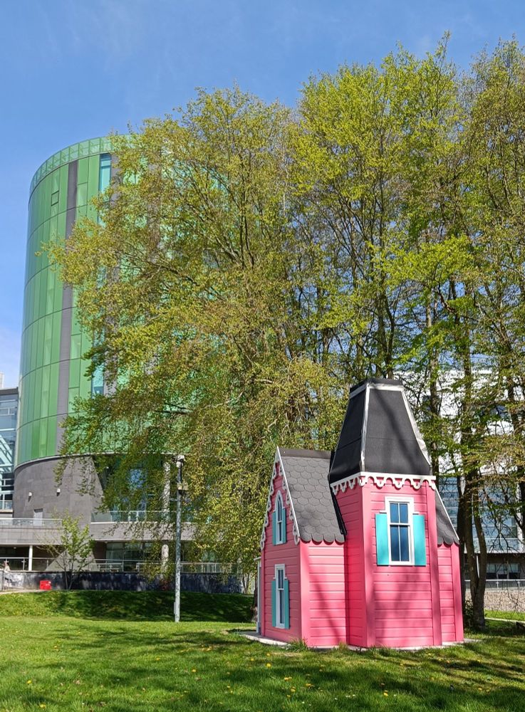 Little pink house artwork in front of the green glass library tour, with a lovely green tree in-between and a perfect blue sky behind it all