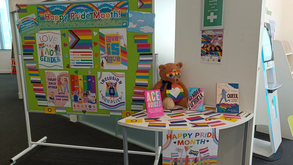 Big notice board of Pride posters and glorious colourful flags next to a table with some relevant books and a teddy bear dressed up like a Pride Care Bear with a rainbow tummy with a lovely heart in the middle 