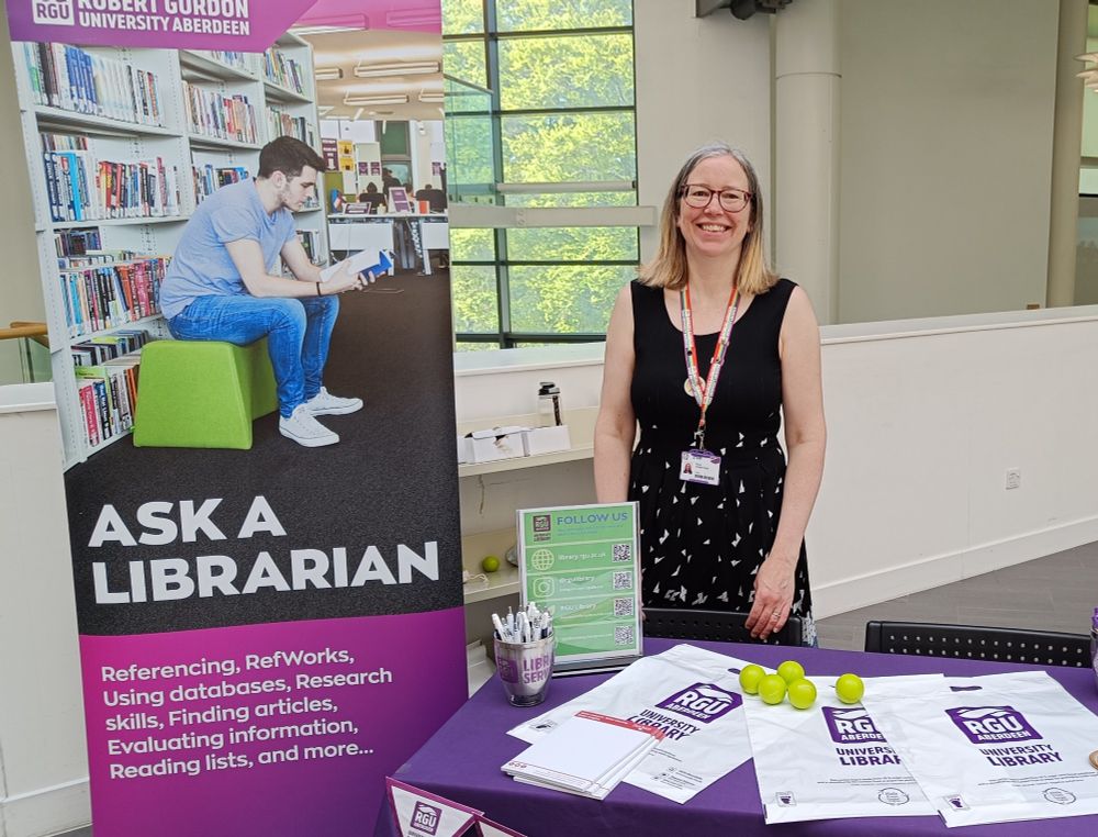 Smily Librarian at a stall with free pens and sugar cane bags with RGU Library logo 
