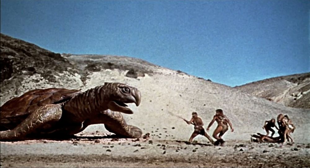 Loana (Raquel Welch) and her seaside tribe rescue a passed out Tumak (John Richardson) from a giant sea turtle. They are on a sandy beach and dunes are in the background. The sky is a pale blue.
