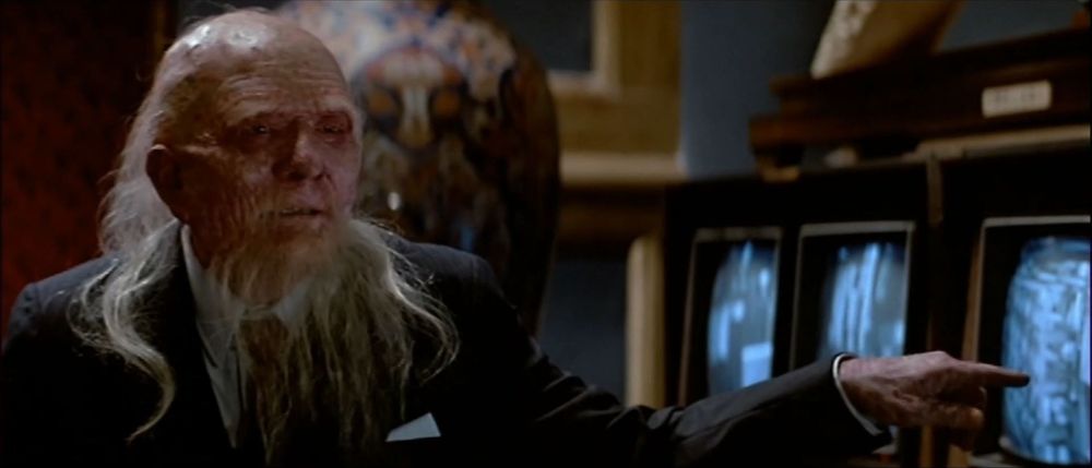 David Lo Pan (James Hong) questions Jack Burton and Wang Chi in his private rooms. He points at monitors showing video feed from the security cameras of their friends snooping around at the front desk of his business. Lo Pan is in a motorized wheel chair, is wearing a dark business suit and has long tuffs of hair growing out of the back of his head and has a long wispy beard and moustache. 