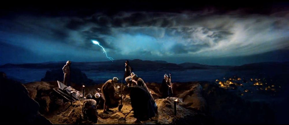 Indy (Harrison Ford), Sallah (John Rhys-Davies) and the hired crew work to uncover the lid that covers the opening to the Well of Souls. A fierce storm of supernatural proportions brews in the background as lightning strikes. An encampment lit by campfires can be seen in the valley at night. 
