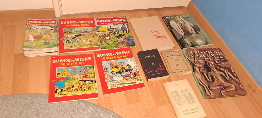 A various assortment of books, roughly from left to right:
- A handful of bright red books called "Suske en Wiske", a very nostalgic comic series written by Willy Vandersteen
- A copy of J.R.R. Tolkien's "The Hobbit"
- A large white book of fairy tales & myths in Lapland
- Two smaller books featuring adventures of Sherlock Holmes
- A book regarding indian mythology
- A book regarding celtic mythology