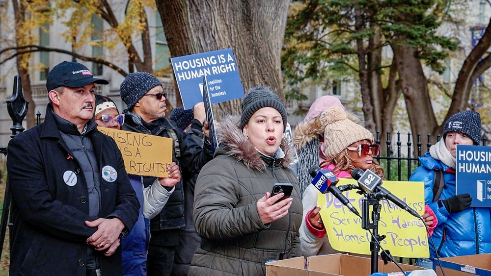 Deputy Public Advocate Gonzalez, dressed for the cold, speaks forcefully at a rally outside of City Hall. Advocates hold posters with messages like "Housing is a human right"