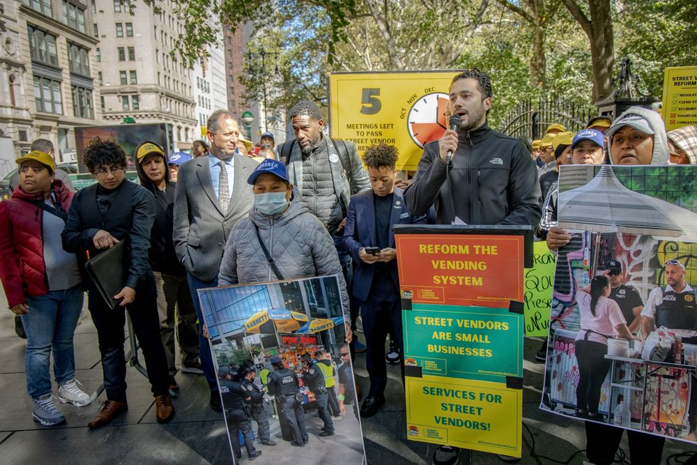 Mo Attia speaks as elected officials and advocates listen, some holding large photos of street vendors interacting with law enforcement