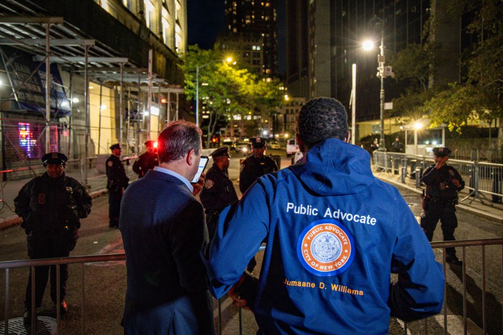 The Public Advocate and Comptroller, photographed from behind, look on past the barricades in front of 26 Federal Plaza. NYPD Officers are on the other side