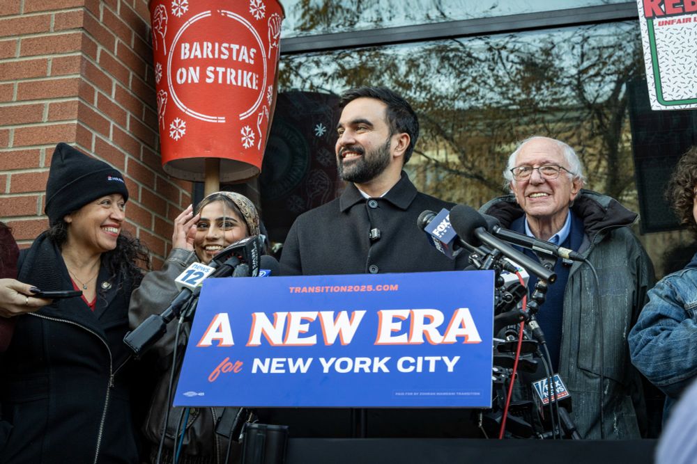 Mayor-Elect Mamdani speaks as Senator Sanders, Council Member Avilés, and advocates smile. A red cup sign reads, "Baristas on strike"