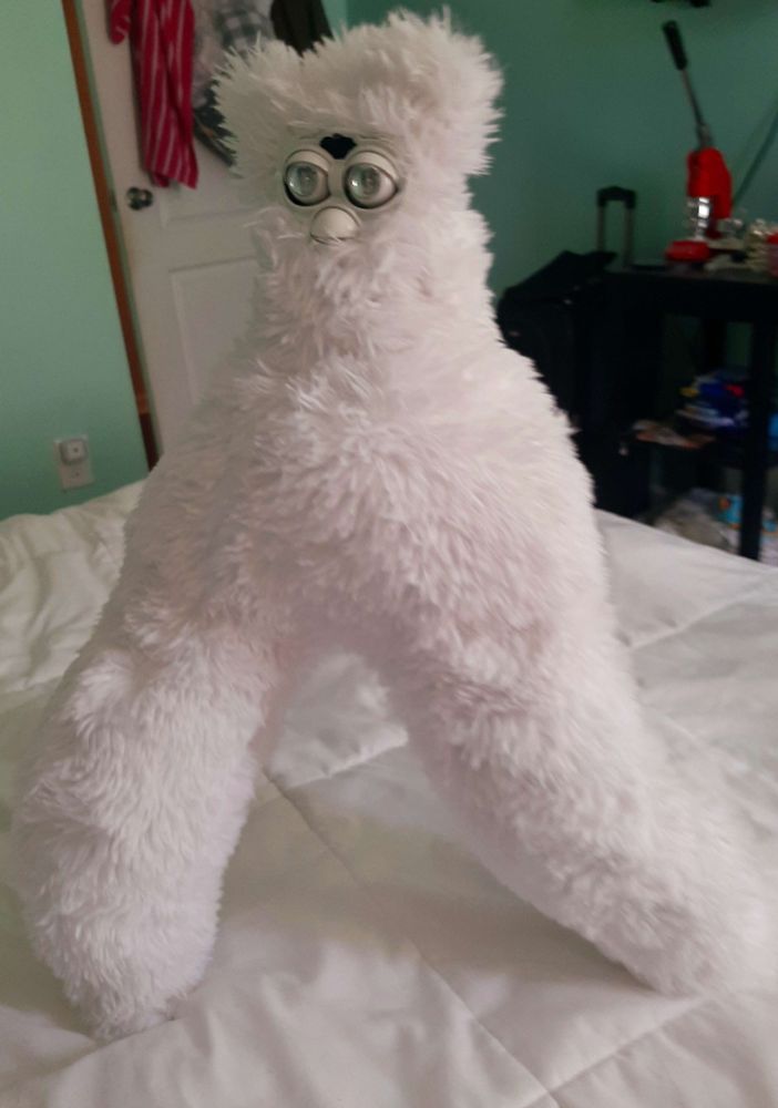 A fluffy white furby with two long legs. Their eyes and clear and glassy and they stride across a bed. 