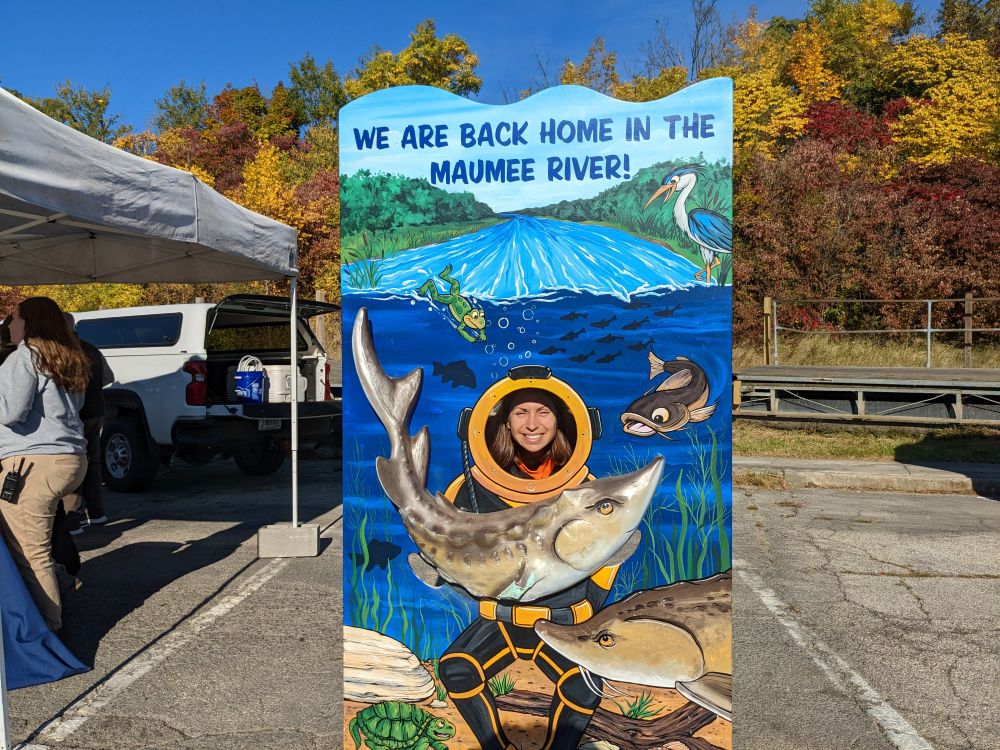 A person stands by a sign that says "We are back home in the Maumee River"