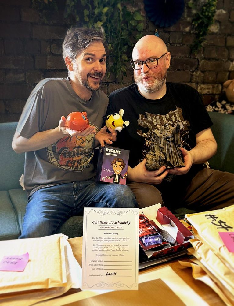 Simon and Lewis sit smiling. They're holding up pieces of Yogscast memorabilia to the camera. There's a 'Certificate of Authenticity' in the foreground.