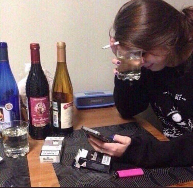 A girl scrolling on her phone at a table with a cigarette in hand. There are 3 bottles of wine on the table.