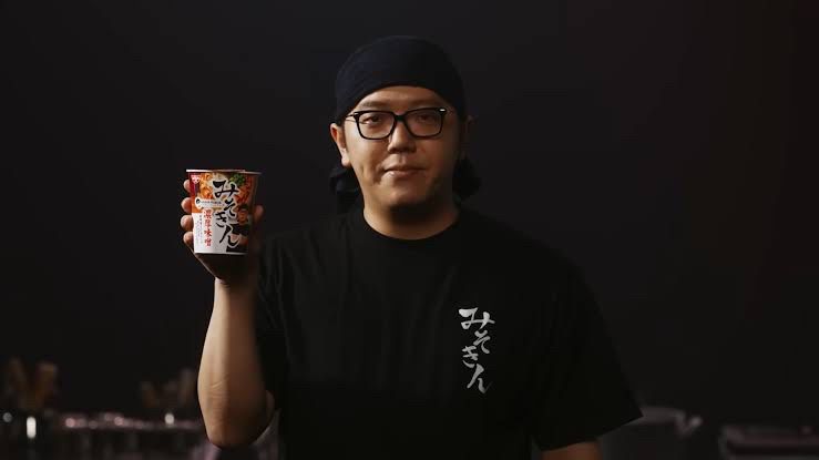 男性が「みそきん」と縦書きされた黒いシャツを着て、緊張した表情でこちらを見ています。
右手には「みそきん」と書かれたカップを持っています。