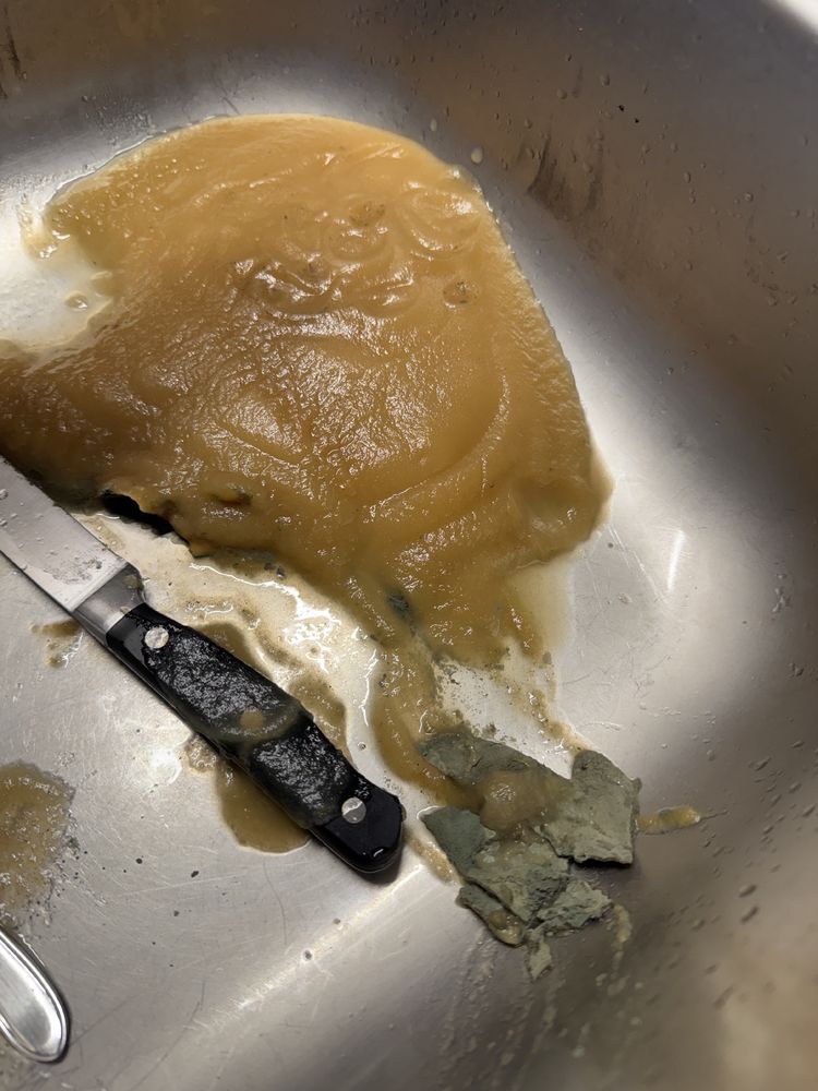 Verdorbenes Apfelmus in einer Edelstahlspüle. Das Apfelmus ist teilweise gräulich und fest, mit schimmelähnlichen Stücken. Ein Küchenmesser mit schwarzen Griff liegt daneben und ist mit Rückständen des Apfelmus bedeckt.