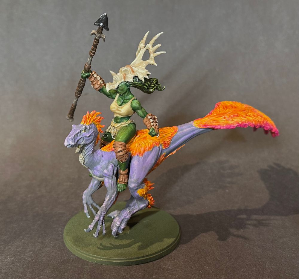 A female orc wearing a triceratops skull on her head, holding up a spear while riding a velociraptor. The raptor has purple skin and orange to magenta feathers