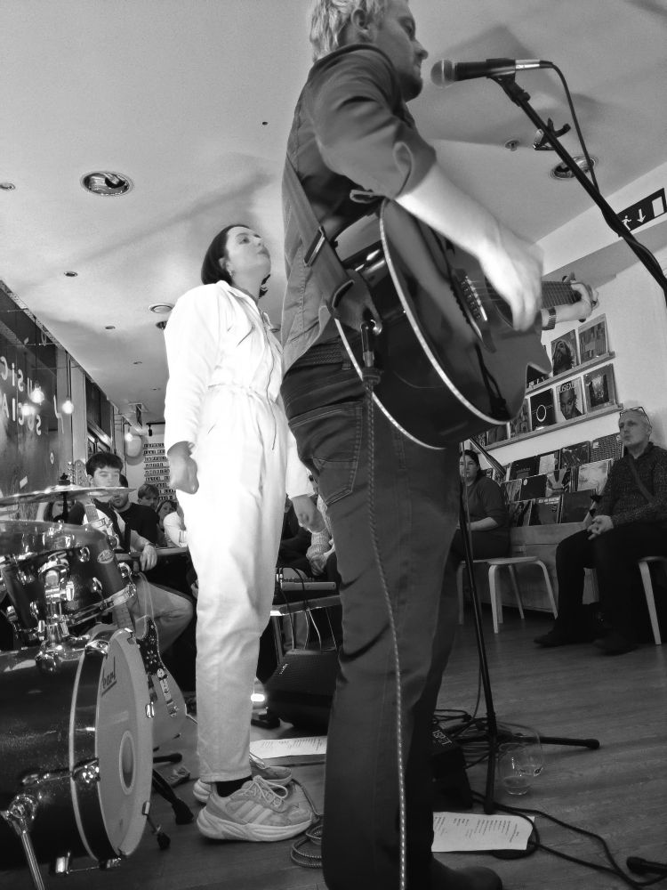 Black and white image of the singer and guitarist of Leonian Dream on stage in Exeter. She (singer) wears a white boiler suit, he (guitarist) sings into a microphone