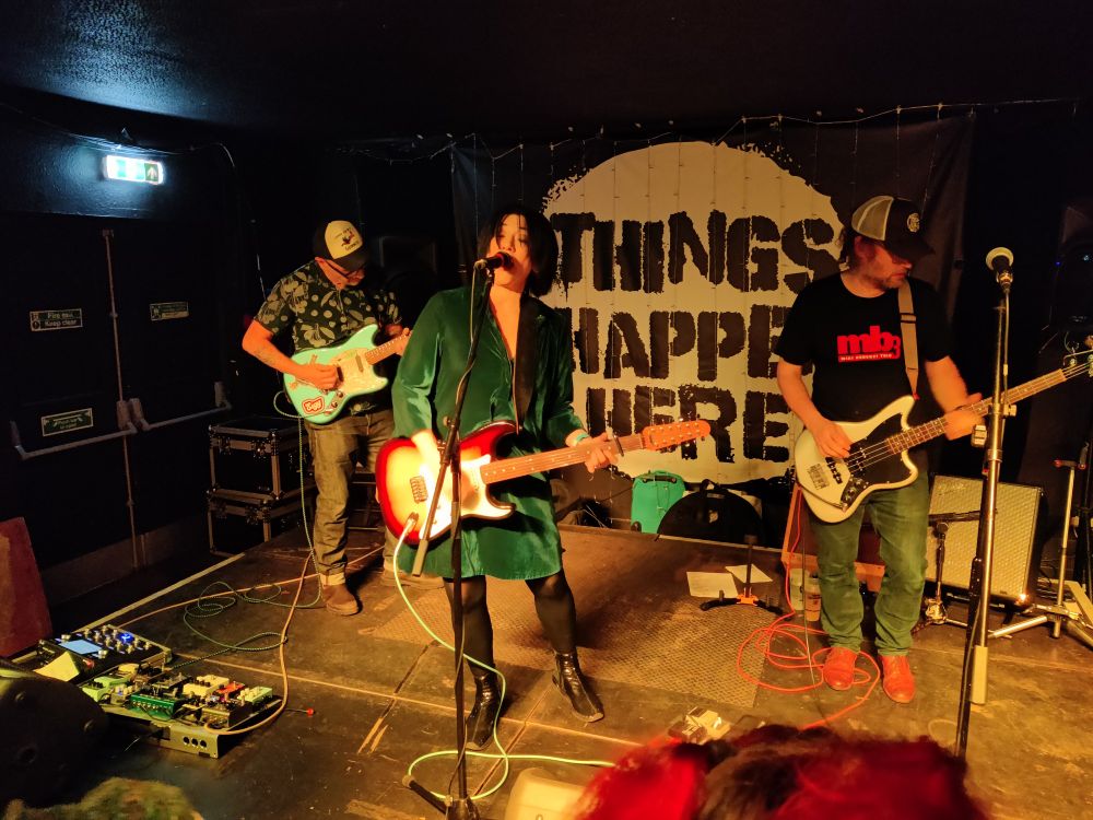 Miki Berenyi Trio on stage with a large backdrop of black text on a white circle saying "Things Happen Here" (the name of the venue in Dartington, Devon).