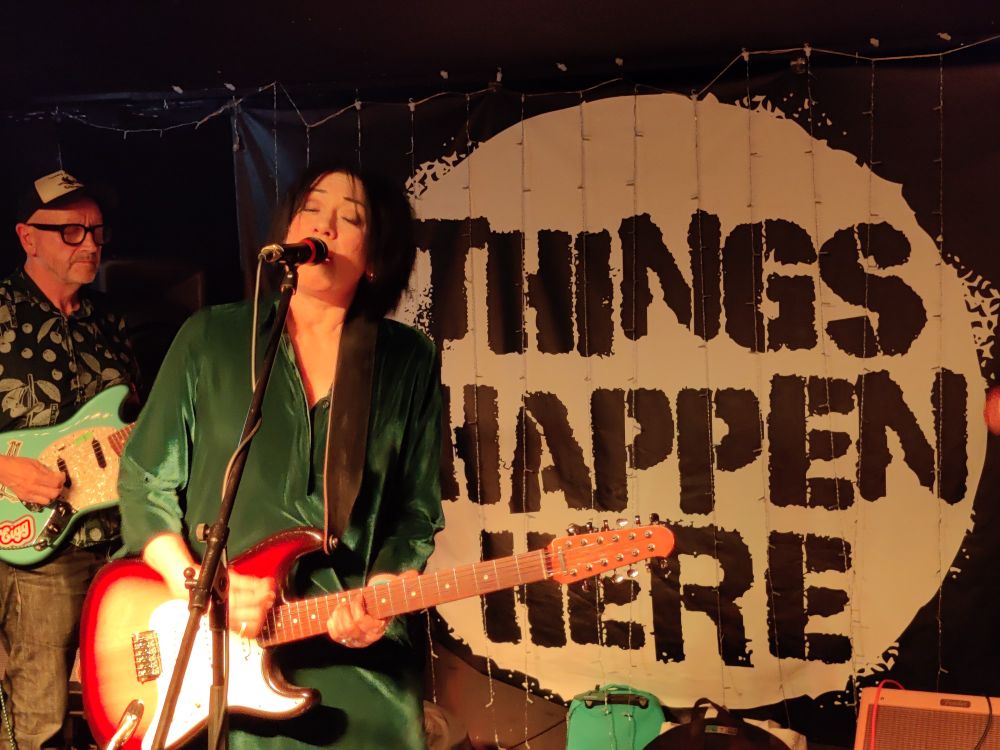 Miki Berenyi on stage at Things Happen Here in Dartington in Devon, wearing a green velvet dress, playing electric guitar and singing into a microphone.