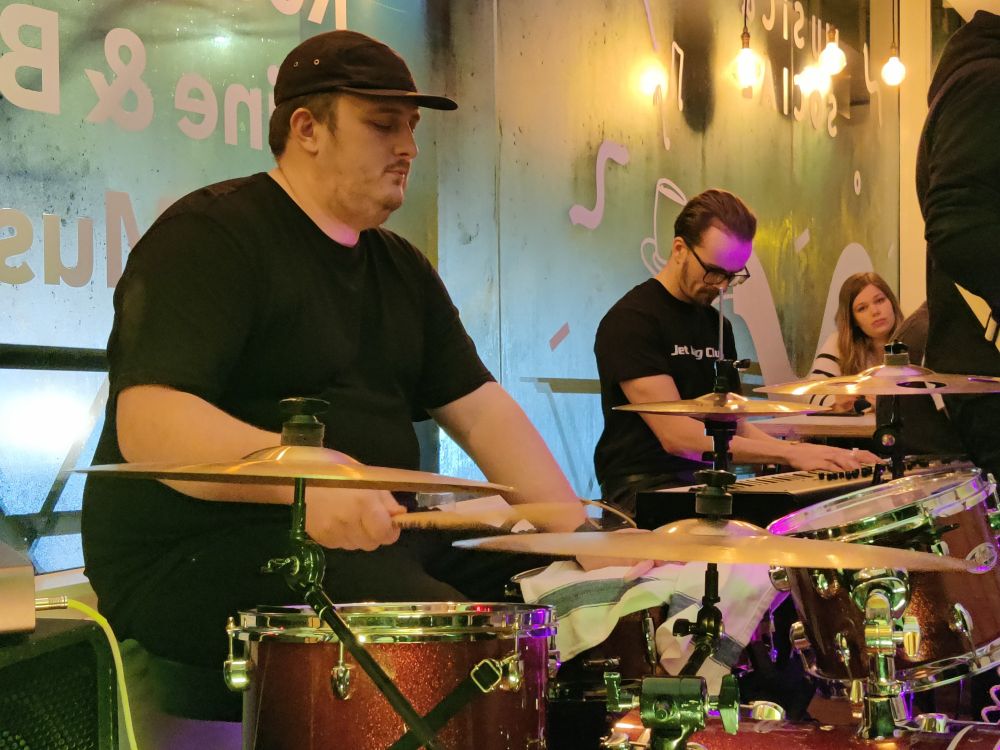 The drummer and keyboard player of Leonian Dream on stage in Exeter. Both are wearing black t-shirts and the drummer wears a black cap. The backdrop is steamy windows.