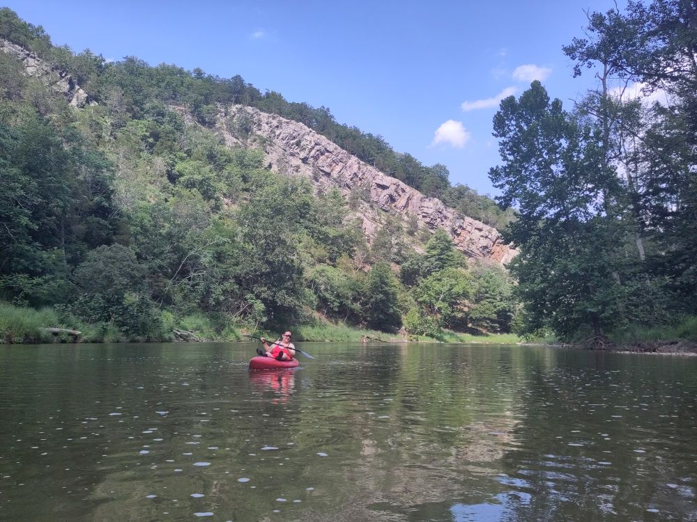 A red kayak with person in it sits in the middle of the river. Above is a long rock wall on top of a mountain. Greenery on the sides and a blue sky above