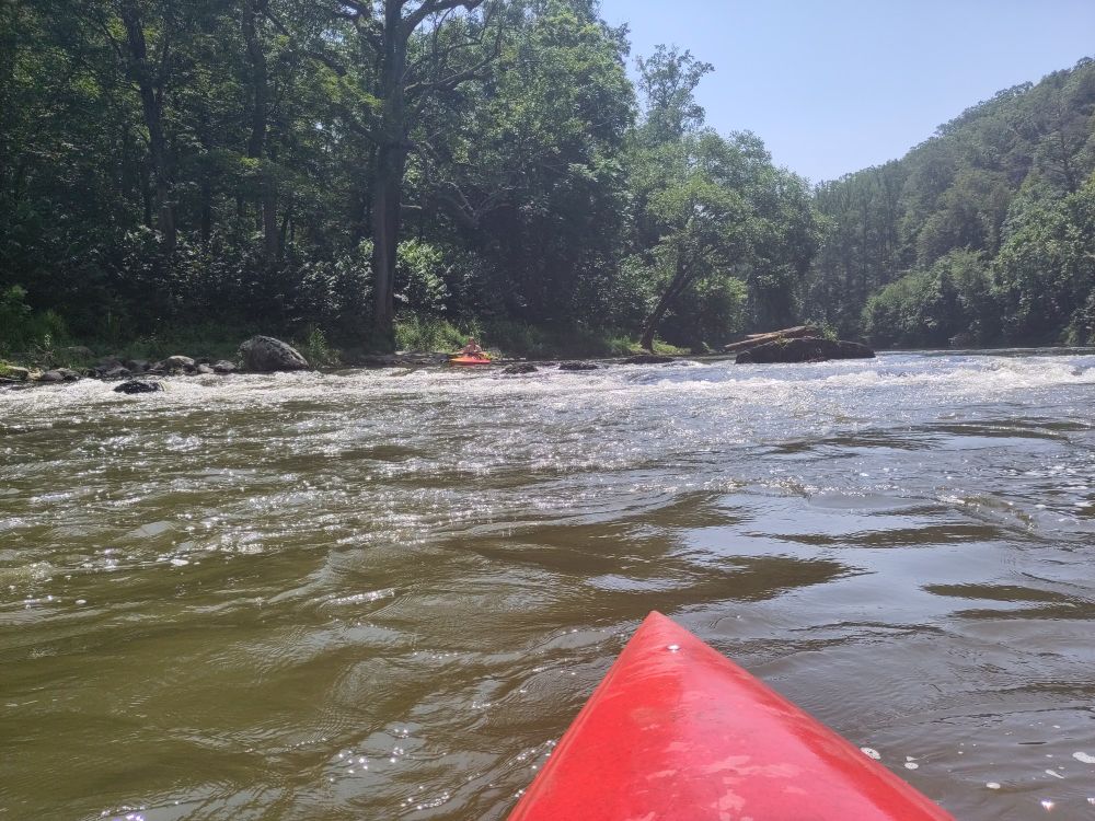 River screen. The red top of a kayak in front. Another kayaker is coming down a small rapid in the distance