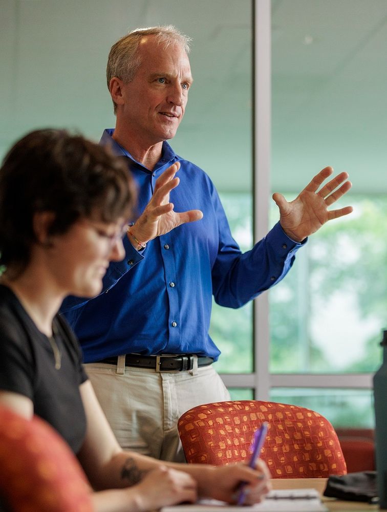 A photo of Dr. Tim Stretton teaching in a classroom at Saint Mary's University. He is wearing a blue shirt and is gesturing while he speaks to students. One student is visible on the left side of the photo, writing in a notebook. 