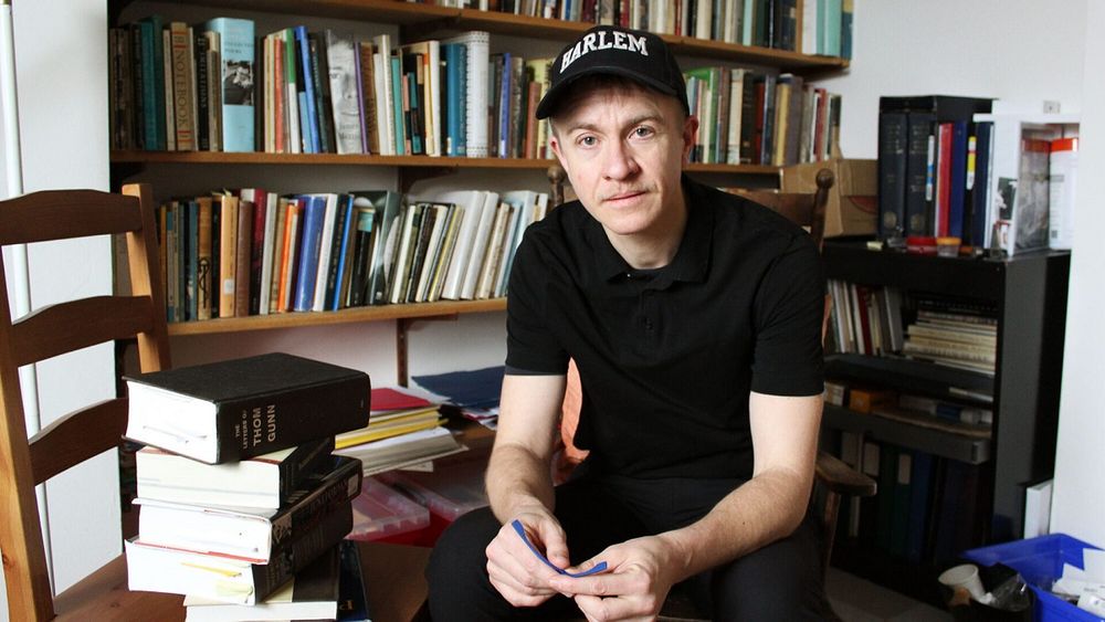 A photo of Dr. Luke Hathaway in his office. Surrounded by books, he is wearing a black t-shirt and black baseball cap. 

Photo by Laura Flight of The Signal, a student publication of the University of King's College School of Journalism in Kjipuktuk/ Halifax. 