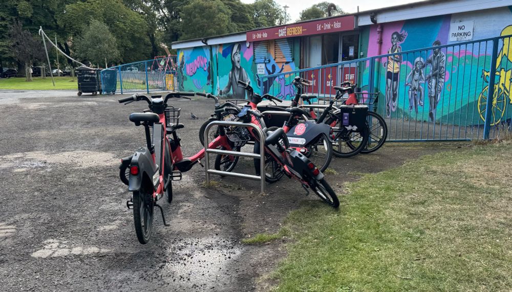 A row of three metal cycle racks is ensconced by a smattering of Coral-coloured Voi hire ebikes; two are parked parallel to the racks like a normal cycle, one is parked upright on its stand nearby, and two are abandoned at a jaunty angle. The patchy asphalt is wet from a recent rainshower; in the background, a colourful mural depicting sportspeople adorns the wall of a single storey building. 
