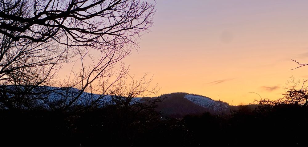 Sunset over a snow-capped Pentland hills - a foreground of bare tree branches and bushes sits silhouetted against a bright, orange-yellow horizon and the white icy caps of the hills ahead.