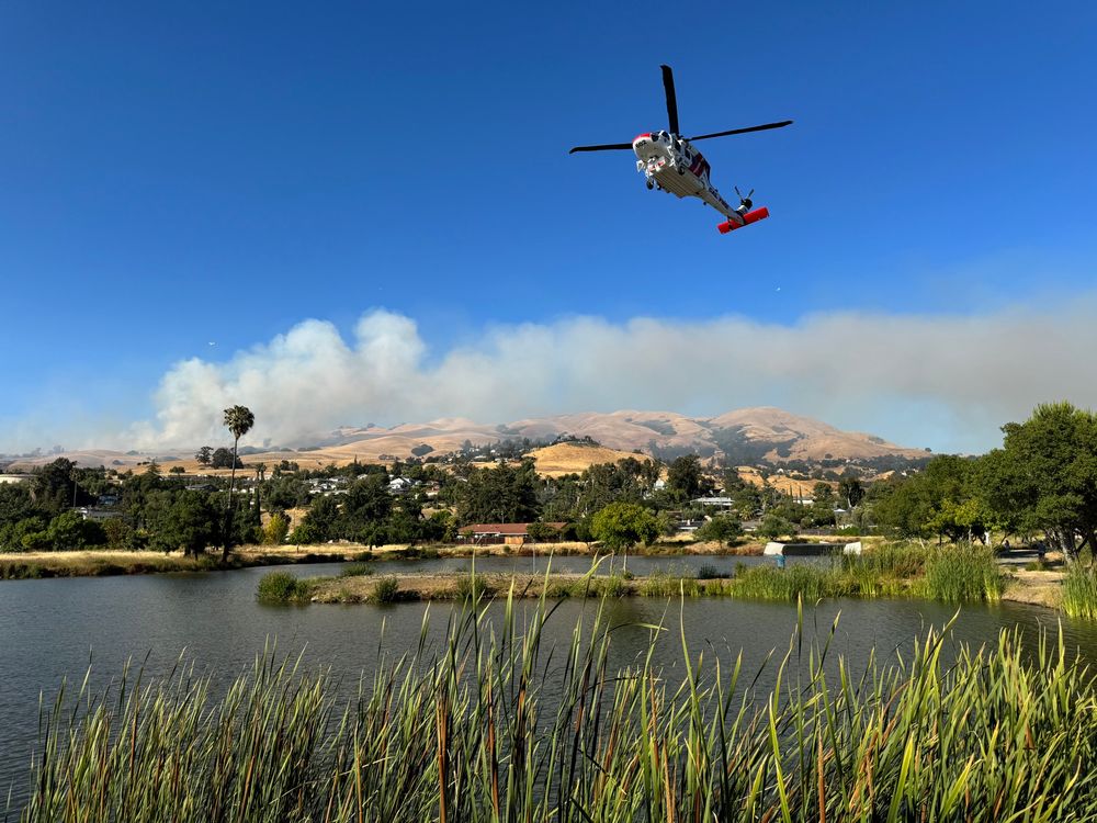 A helicopter approaching a pond with smoke and hills in the background